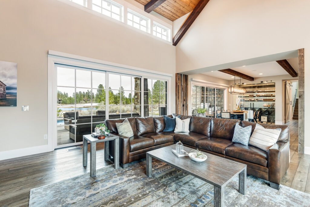Modern rustic living room with lake views, leather sectional sofa, geometric rug, wood beams, and open layout connecting to dining and kitchen areas.