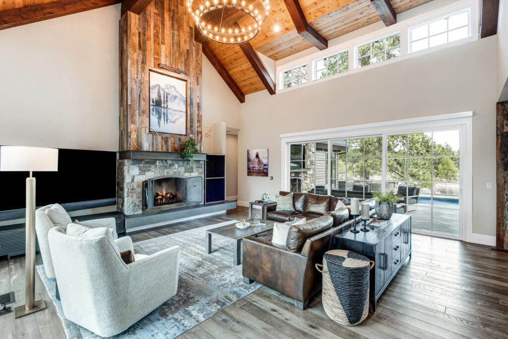 Spacious living room with vaulted wood-beam ceiling, stone fireplace, leather sofa, large windows, and chandelier—blending rustic charm and modern comfort.
