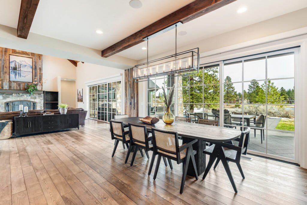 Open-plan rustic modern living and dining area with wooden dining table, woven chairs, stone fireplace, wood beams, and large windows with outdoor views.