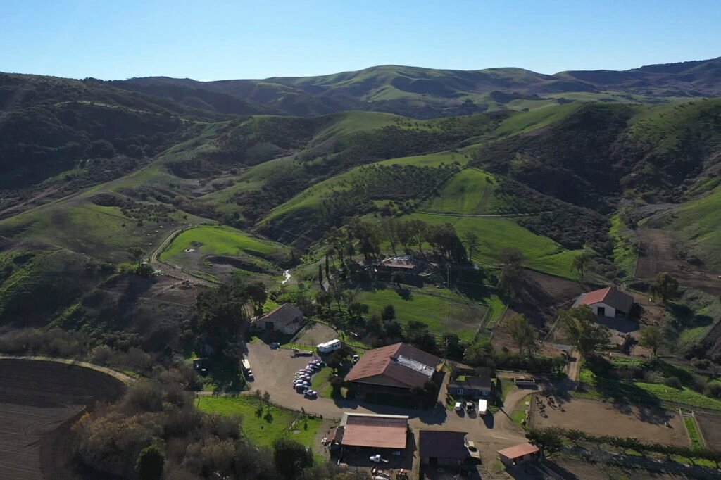 Aerial view of a ranch with buildings and vehicles in a green valley surrounded by rolling hills.