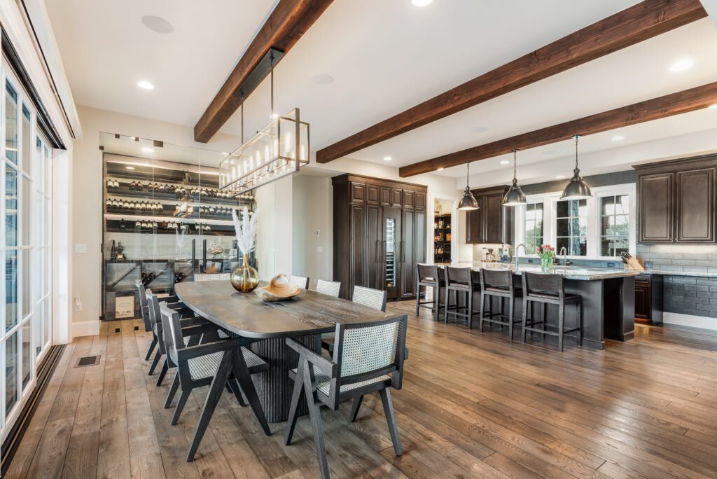 Modern kitchen and dining area with Rivièra Maison St. Moritz armchairs, dark wood table, wine storage, wood beams, and natural light in a luxury home.