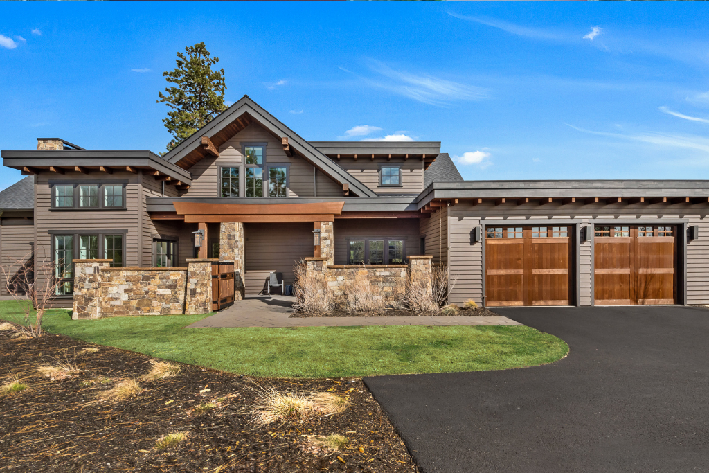 Exterior of modern home at 61582 Searcy Ct, Bend, OR, featuring wood siding, stone accents, two-car garage, landscaped yard, and clear skies in a natural setting.