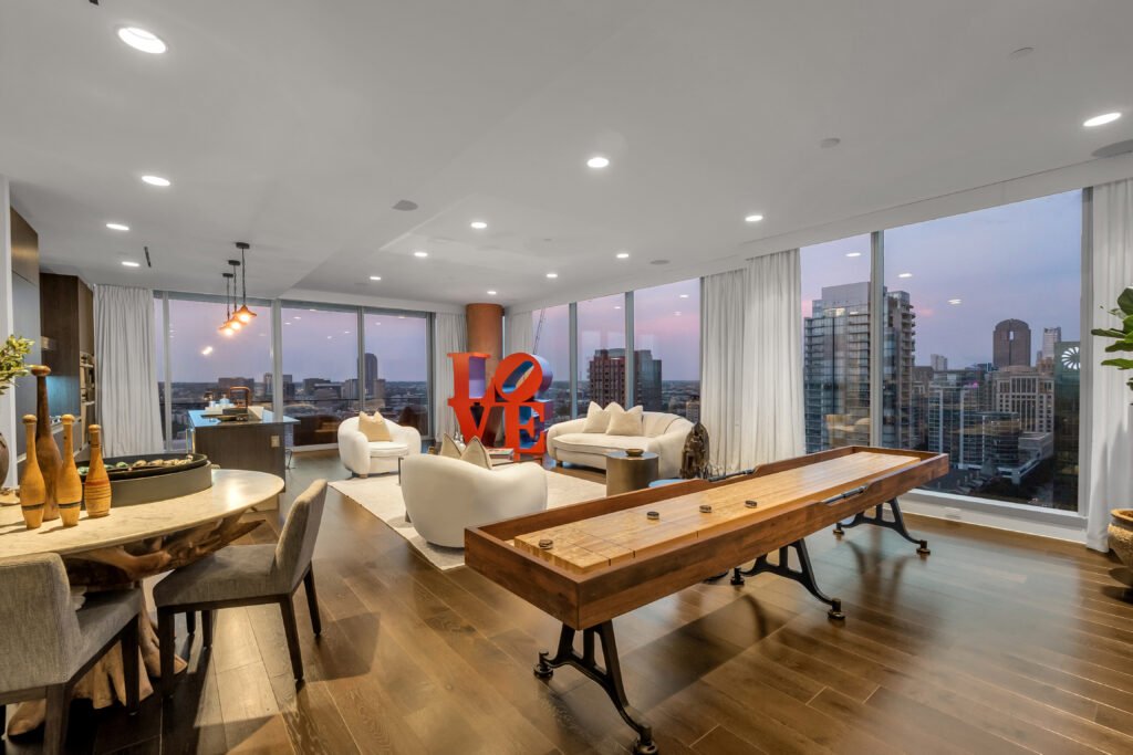 Modern penthouse living room with city skyline views, hardwood floors, contemporary furniture, shuffleboard table, and vibrant "LOVE" sculpture—urban luxury in Dallas.