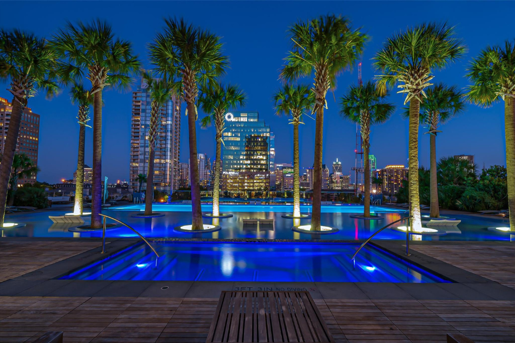 Bleu Ciel Dallas at night with illuminated blue pool, palm trees, and city skyline—showcasing modern architecture and luxury high-rise amenities.