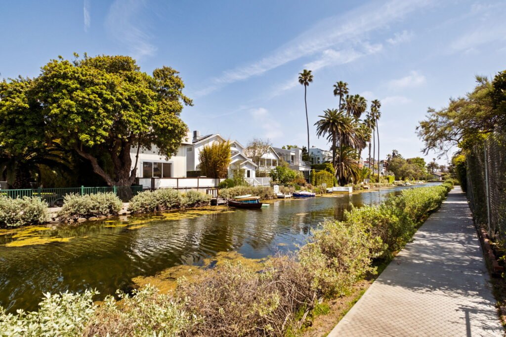 A scenic view of the Venice Canals in Venice, CA, featuring a row of modern and charming homes along the water, small docked boats, lush greenery, tall palm trees, and a walking path under a clear blue sky.