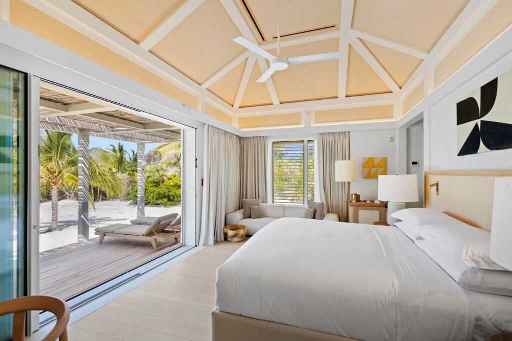 Luxurious bedroom featuring a large white bed beneath a vaulted ceiling with a ceiling fan, and a wide glass sliding door opening onto a private patio with lounge chairs, overlooking a sandy beach with palm trees and the ocean in the background.