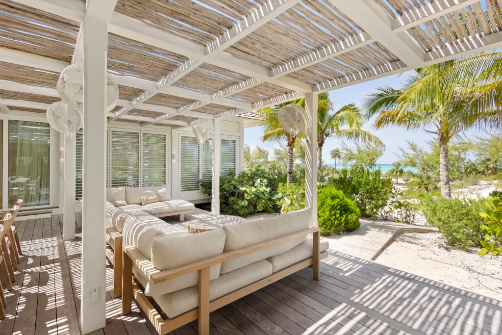 Bright outdoor living space with a large light-colored sofa and lounge chairs under a white pergola with a straw roof, set on a wooden deck beside a sandy beach with palm trees, lush greenery, and ocean views in the distance, featuring a Piet Boon Lars daybed and Chrysobalanus icaco plants.