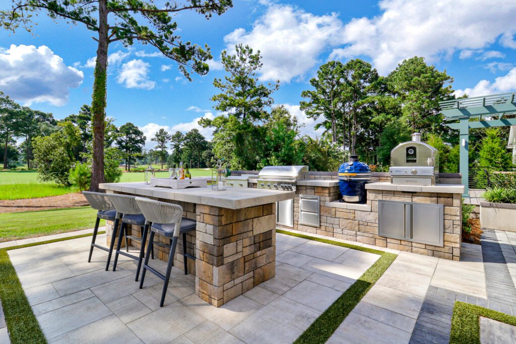 : Outdoor kitchen with a sleek high-top bar and cushioned seating, equipped with a 40-inch built-in gas grill, gas griddle, stainless steel oven, and ceramic smoker, blending style and functionality.