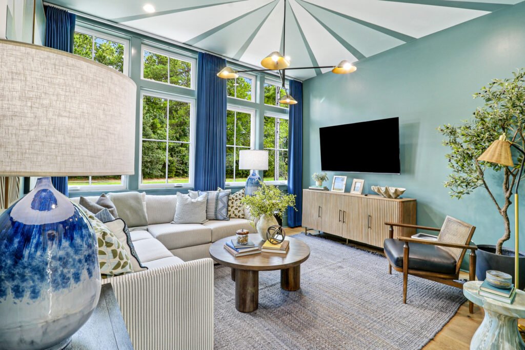Bright, modern living room with large windows dressed in blue curtains, light-colored sectional sofa with decorative pillows, round wooden coffee table, dark woven-back armchair, and flat-screen TV mounted on a light blue wall above a wooden console table. 