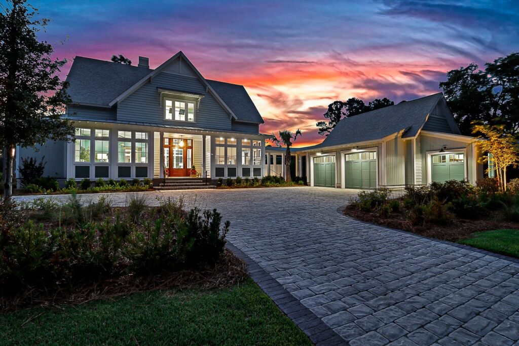 Grand modern Lowcountry-style home, the HGTV Dream Home 2025 in Bluffton, South Carolina, with a main house and detached three-car garage connected by a paved driveway. Captured at sunset with an orange, pink, and purple sky, the scene highlights multiple windows, a welcoming entrance, and meticulously landscaped grounds.