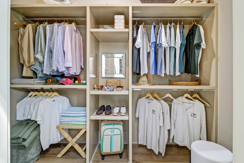 Walk-in closet with sections for hanging clothes, open shelving with a mirror and shoes, folded items, caps, a small suitcase, and a white stool in the foreground.