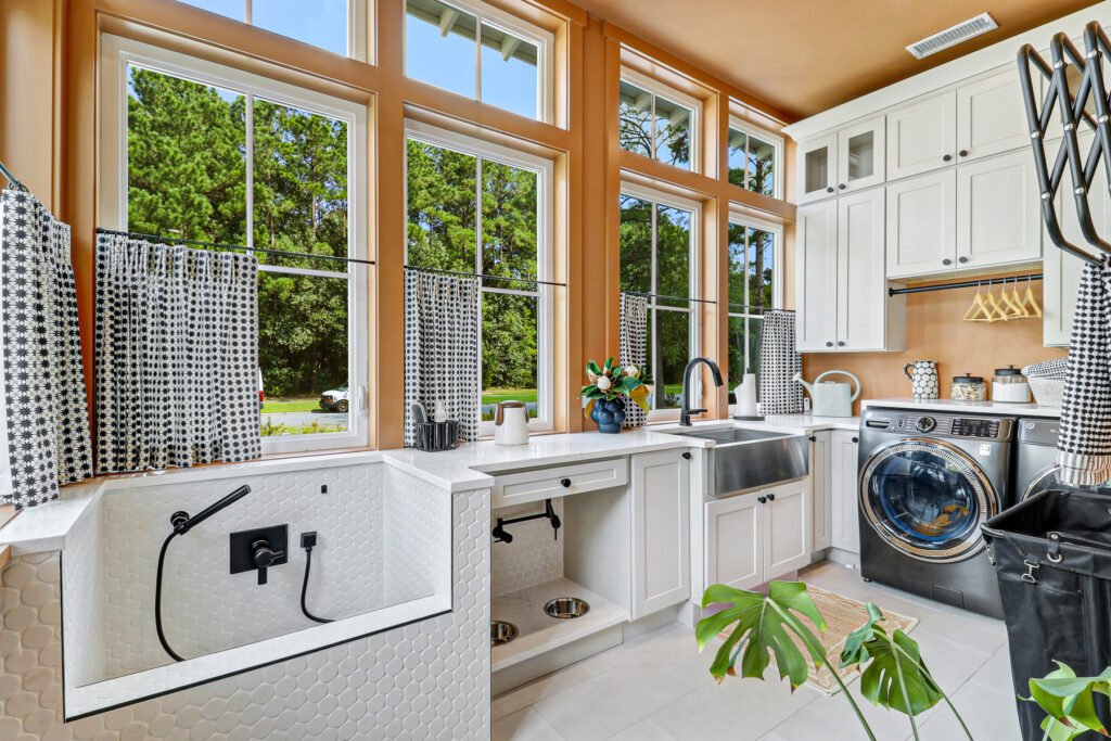 Bright kitchen with white shaker cabinets, light marble countertops and backsplash, stainless steel appliances including a double-drawer dishwasher, large window above a sink with pull-down faucet, open shelves holding baskets and jars, and light blue painted walls.