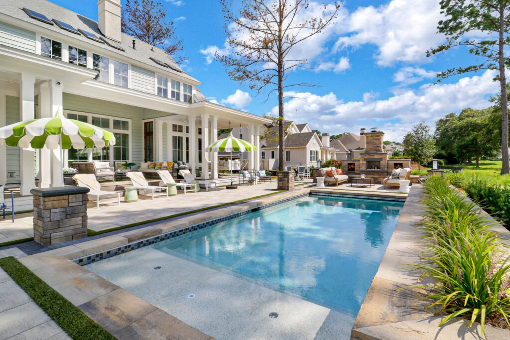 Luxurious backyard at 3500 Holly Road, Virginia Beach, VA, featuring a large light-blue swimming pool, stone patio with lounge chairs, green and white striped umbrellas, outdoor fireplace with seating, lush landscaping, and a view of the light-colored house with covered porch under a partly cloudy sky.