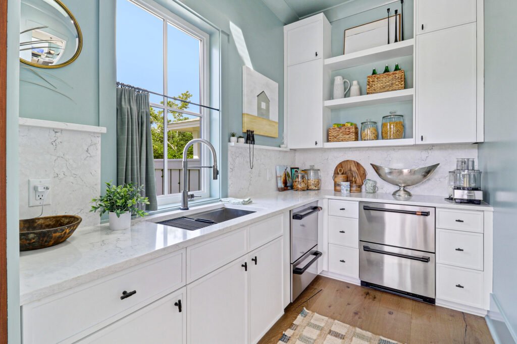 Bright kitchen with white shaker cabinets, light marble countertops and backsplash, stainless steel appliances including a double-drawer dishwasher, large window above sink with pull-down faucet, open shelves holding baskets and jars, and light blue painted walls.