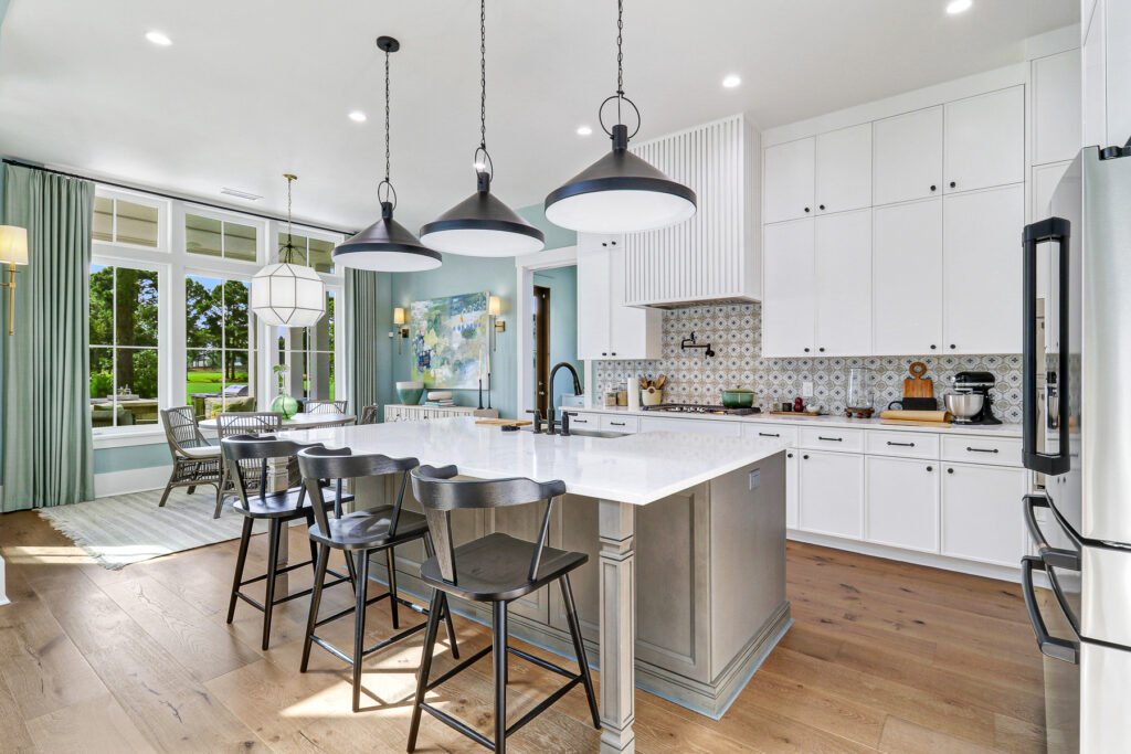 Bright, open-concept kitchen from an HGTV Dream Home with ceiling-high white cabinets, large island with white countertop and gray base, dark counter stools, three pendant lights, decorative tiled backsplash above range, wide-plank wood floors, and adjacent dining area with tranquil green walls visible through large windows.