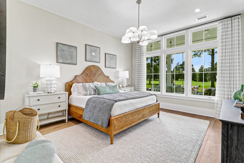 Bright primary bedroom with a wooden herringbone headboard bed, radial-design nightstands, large windows overlooking a golf course, a multi-tiered chandelier, and neutral-toned decor with woven rug and framed artwork.