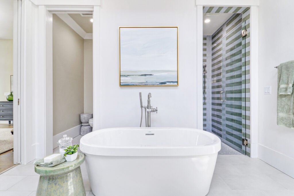 Bright modern bathroom with a white freestanding bathtub and chrome floor-mounted tub filler in the foreground, a glass-enclosed shower with green and white striped tiles on the right, and a doorway to another room on the left. Above the tub hangs a large blue and gray abstract painting, and a green stool with a small plant and glass jar sits beside it.