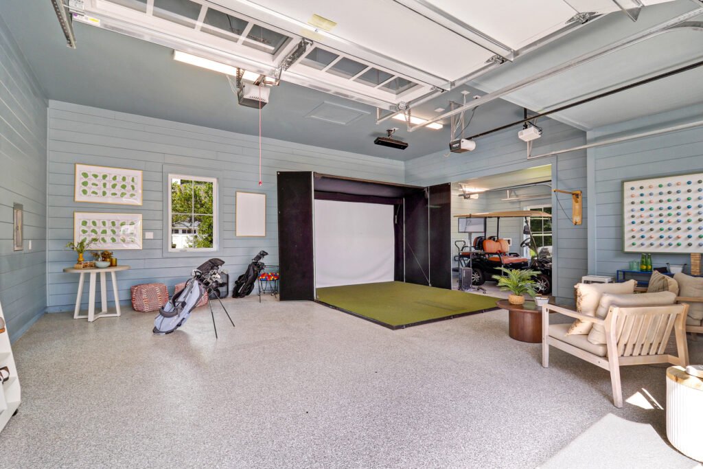 Spacious converted garage recreational area with light epoxy flooring, light blue-green walls, and white trim, featuring a golf simulator with large screen and green mat, a seating area with two armchairs and coffee table, a golf cart in the background, framed wall art, and overhead garage door mechanisms.
