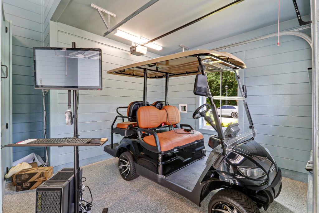 Interior of a garage with speckled grey flooring, featuring a black golf cart with brown seats, a workstation with a large monitor, keyboard, and computer tower to the left, light-colored slatted walls, overhead lighting, and a partially open garage door revealing a vehicle outside.