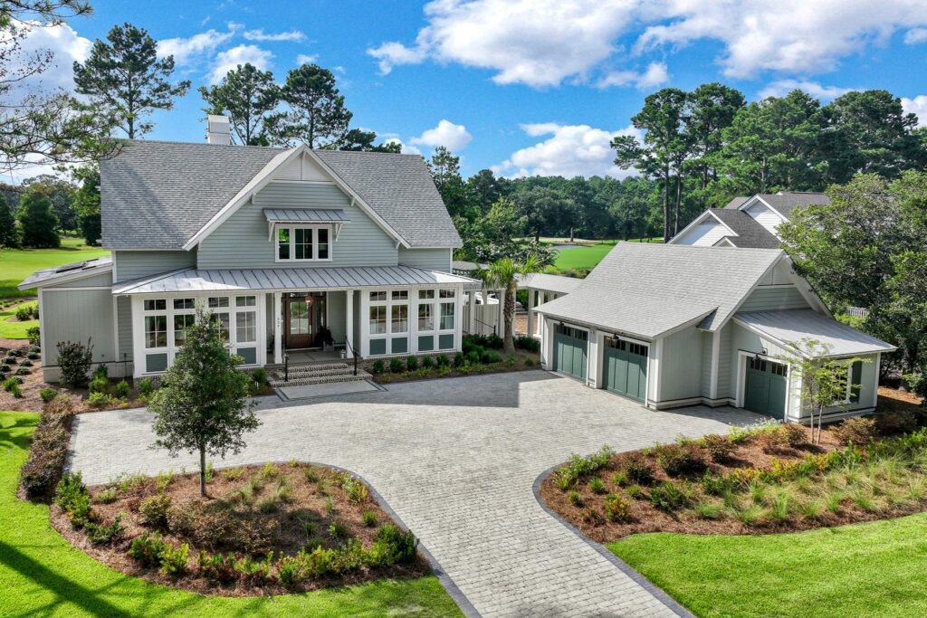 Large light-colored house with grey roof and detached two-car garage with green doors, set on a manicured lawn with a paved driveway, surrounded by trees and a golf course in the background.