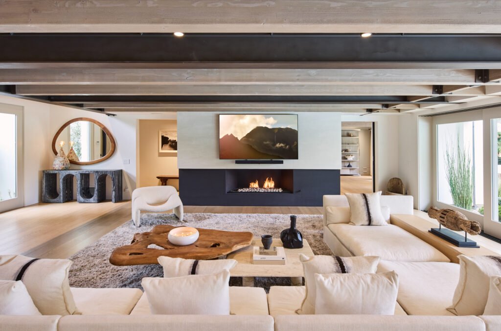A spacious modern living room with a large white sectional sofa, live-edge and marble coffee tables, a fireplace with a mounted TV, exposed wooden ceiling beams, and a textured rug on light flooring.