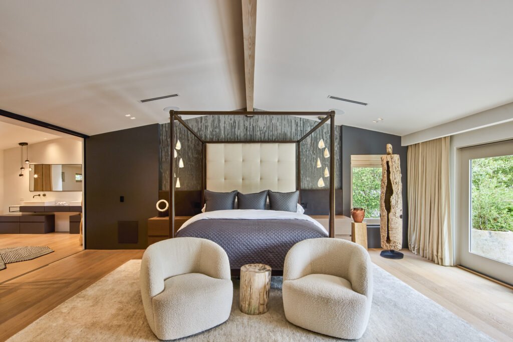 A modern bedroom with a dark four-poster bed, gray bedding, and circular-accent nightstands, featuring two textured armchairs on a plush rug, a doorway to a double-vanity bathroom, and large windows opening to greenery, with high ceilings and exposed wooden beams.