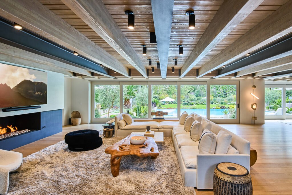 A luxurious contemporary living room with an L-shaped white sofa, wooden coffee table, shaggy rug, and fireplace, featuring beamed ceilings and glass doors opening to a backyard with a pool and greenery.