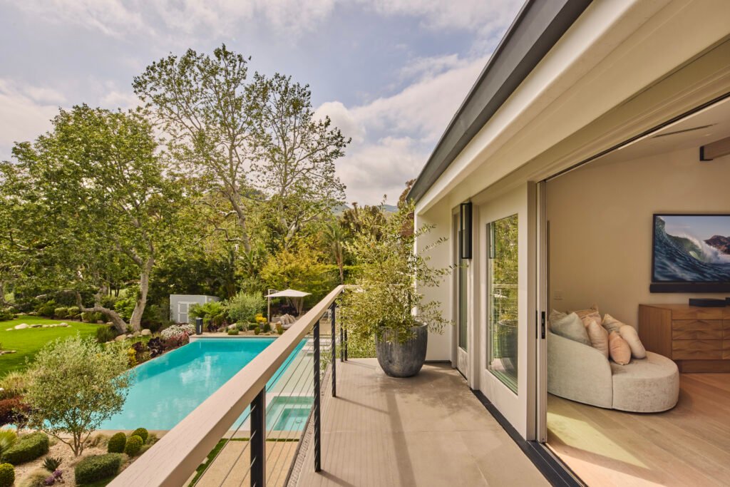 A modern balcony with dark flooring and light railing, overlooking a swimming pool and lush landscaped backyard with mature trees, featuring a large potted plant near sliding glass doors that open to a bright interior with a curved sofa and a TV displaying a wave