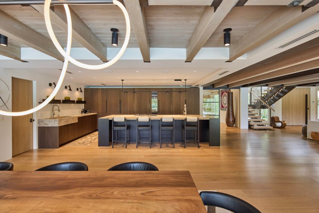 A spacious modern kitchen and dining area with a large wooden island and bar stools, sleek appliances, exposed wooden ceiling beams, and contemporary lighting, with a dining table in the foreground and staircase in the background.