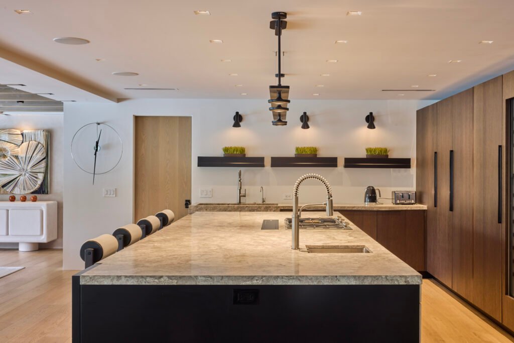 A modern kitchen with a large granite island, dark base and stools, gooseneck faucet, floating shelves with greenery, tall wood cabinets, and light wood flooring.