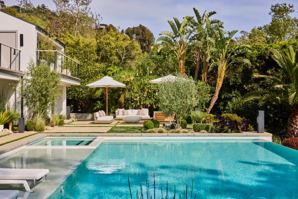 A luxurious Malibu estate backyard with a swimming pool and Baja shelf, lush lawn with seating and umbrellas, mature palm trees, and a modern white house with balcony against a green hillside.