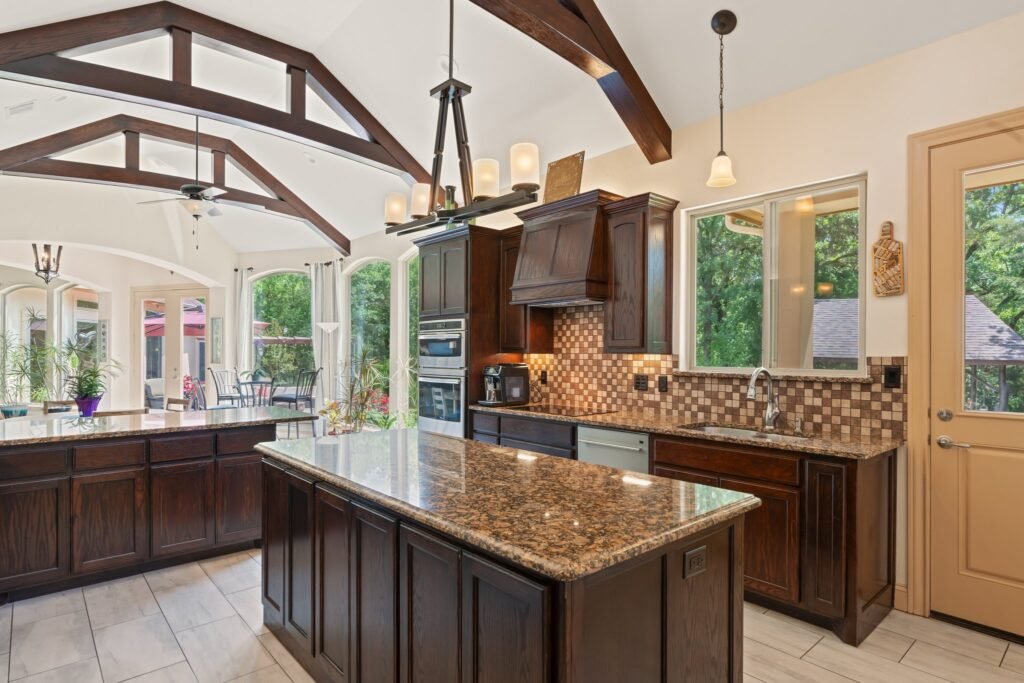 Spacious kitchen with dark wood cabinets, a large granite island with sink, range hood above the stovetop, and appliances, opening into a bright living area with large windows and exposed wooden ceiling beams.