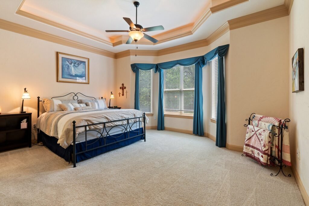 A spacious bedroom with a large bed, ceiling fan, bay window with teal curtains, and light-colored carpet.