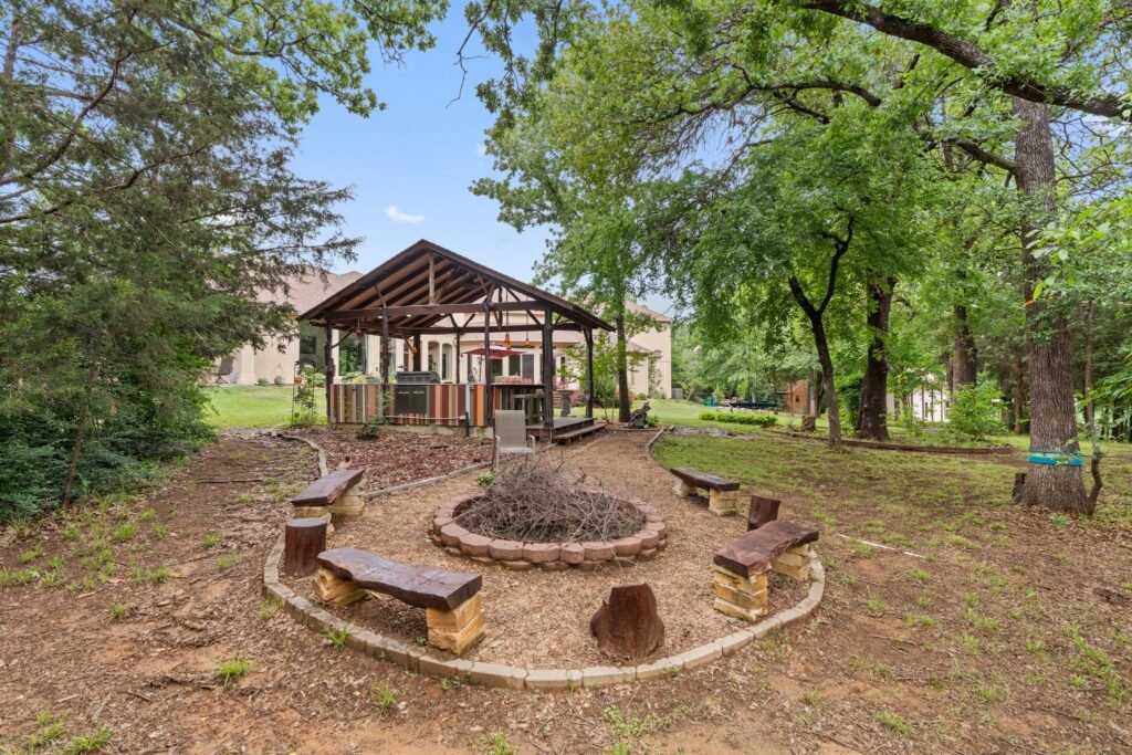 A spacious backyard with a covered outdoor kitchen pavilion, a circular stone fire pit surrounded by benches, and mature trees providing shade in a natural setting