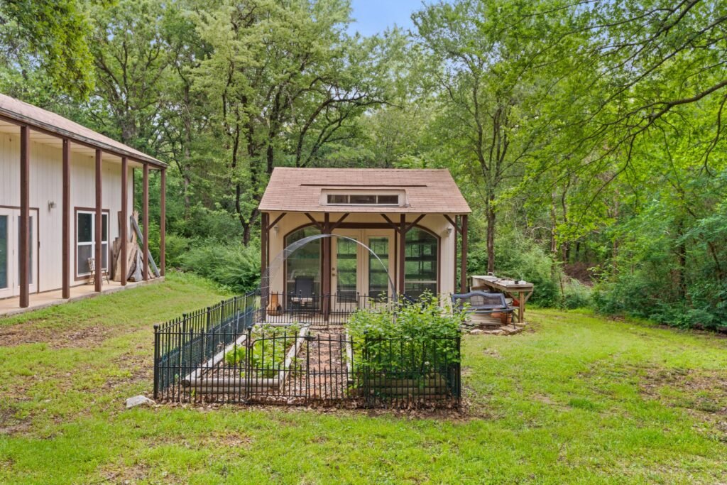 A small shed-like structure with a brown roof and arched windows beside a fenced garden, with trees and part of a larger light-colored building in the background.