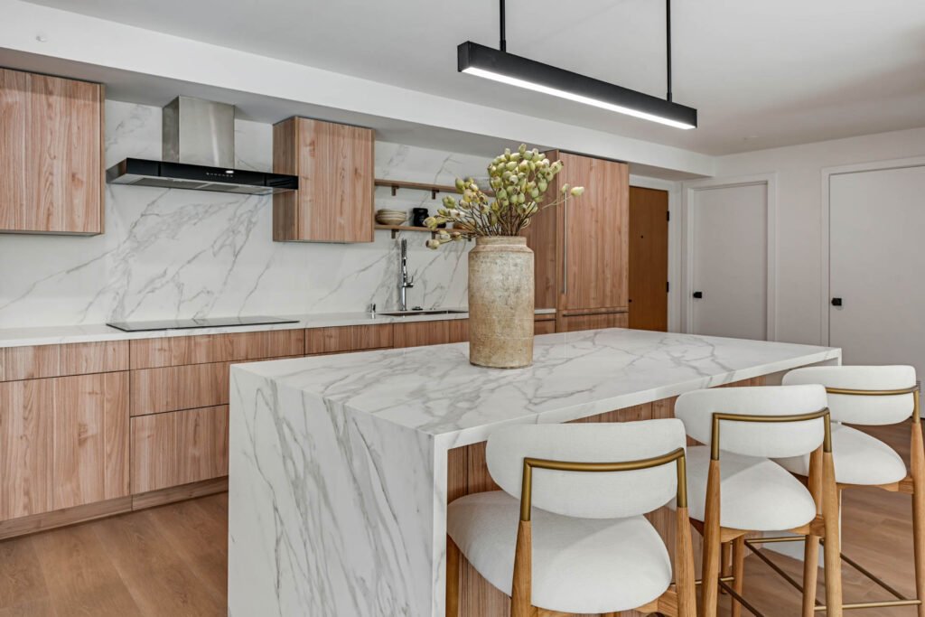Modern kitchen with a white marble waterfall island, three white and gold bar stools, light wood cabinetry, marble backsplash, and stainless steel range hood.