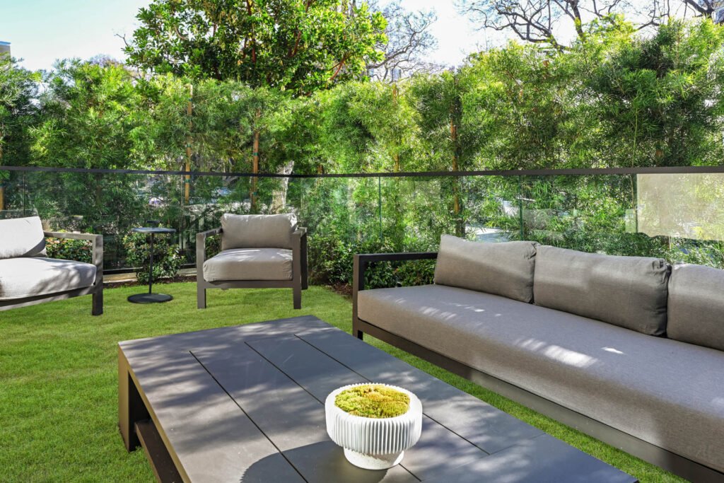 A modern kitchen featuring a large white marble waterfall island with three white and gold bar stools. The kitchen has light wood flat-panel cabinetry, a white marble backsplash and countertops, and a stainless steel range hood above a stovetop. A vase with dried flowers sits on the island, and a long black linear light fixture hangs above it. 