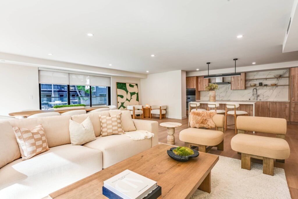 Bright open-concept living room and kitchen with a cream L-shaped sofa, wooden coffee table, armchairs with ottomans, and a modern kitchen with light wood cabinetry, white countertops, and bar seating, lit by large windows with blinds and accented by green abstract artwork.