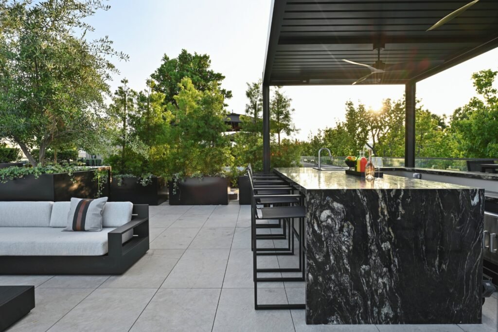 A modern kitchen featuring a large white marble waterfall island with three white and gold bar stools. The kitchen has light wood flat-panel cabinetry, a white marble backsplash and countertops, and a stainless steel range hood above a stovetop. A vase with dried flowers sits on the island, and a long black linear light fixture hangs above it. 