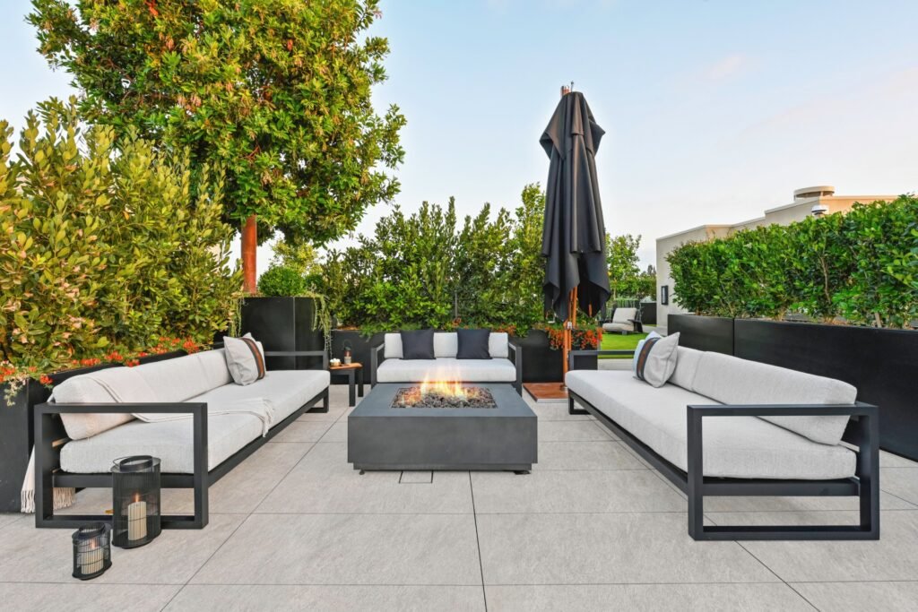 "Luxurious rooftop terrace at Palma Beverly Hills with modern outdoor sofas around a rectangular fire pit, lush greenery in planters, a large black patio umbrella, and lanterns with candles on a tiled floor.