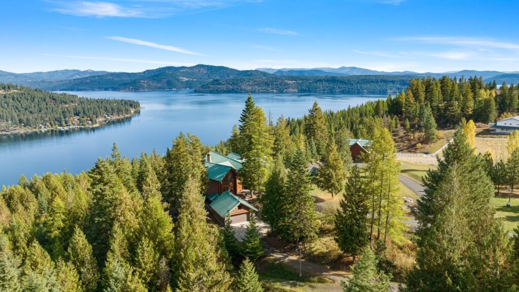 Aerial view of a large log cabin with a green roof, surrounded by evergreen trees on a hillside overlooking a lake and distant mountains under a clear blue sky.