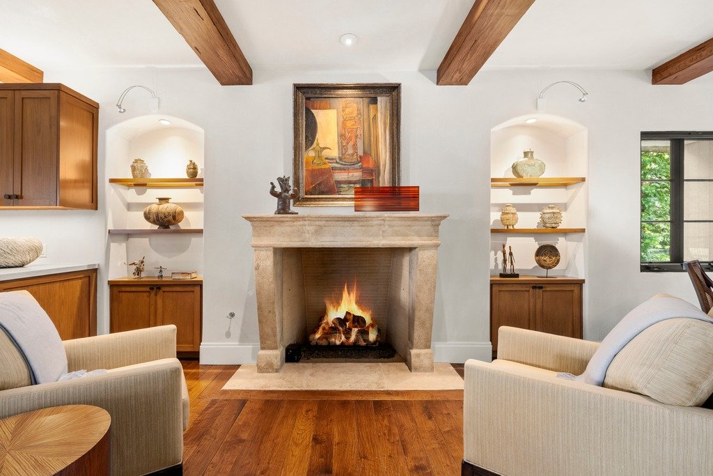 Mediterranean-style living space with a large stone fireplace, flanked by built-in wooden shelves displaying decorative objects, dark ceiling beams, warm wood flooring, and two light-colored armchairs facing the fire, with a framed artwork above the mantel and a window overlooking greenery.