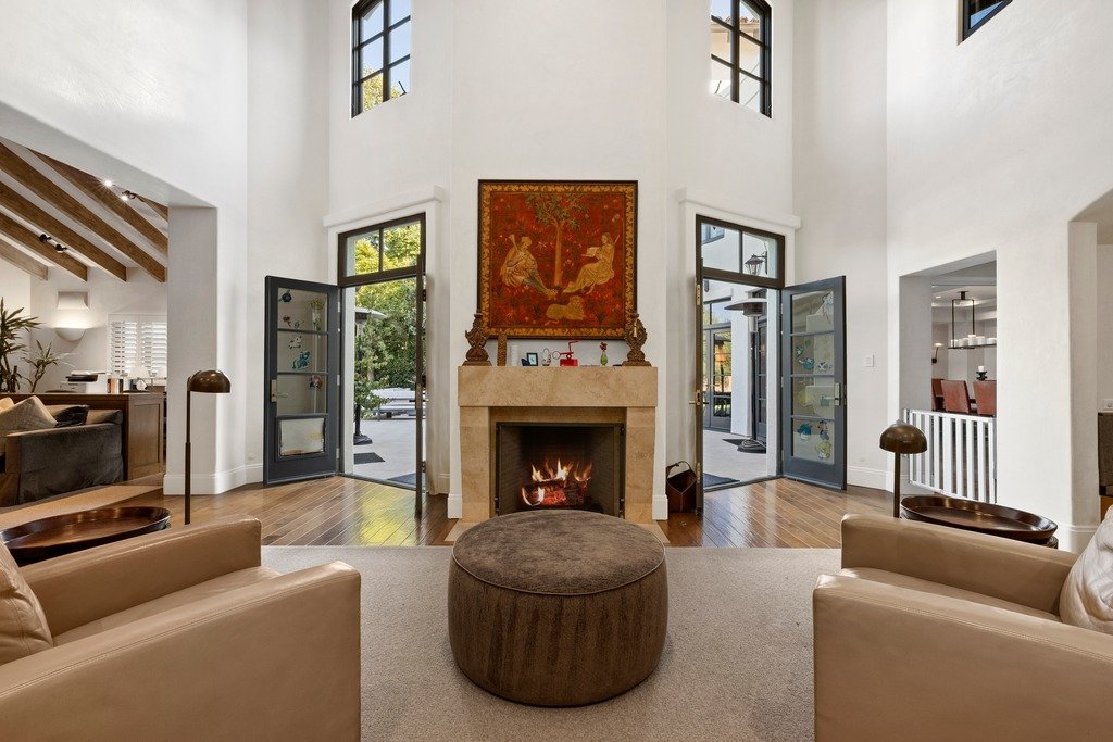 Luxurious multi-level Spanish-style estate at dusk with warm interior lighting, arched doorways, dark-framed windows, terracotta tile roof, and a wide driveway, surrounded by lush trees and manicured landscaping.