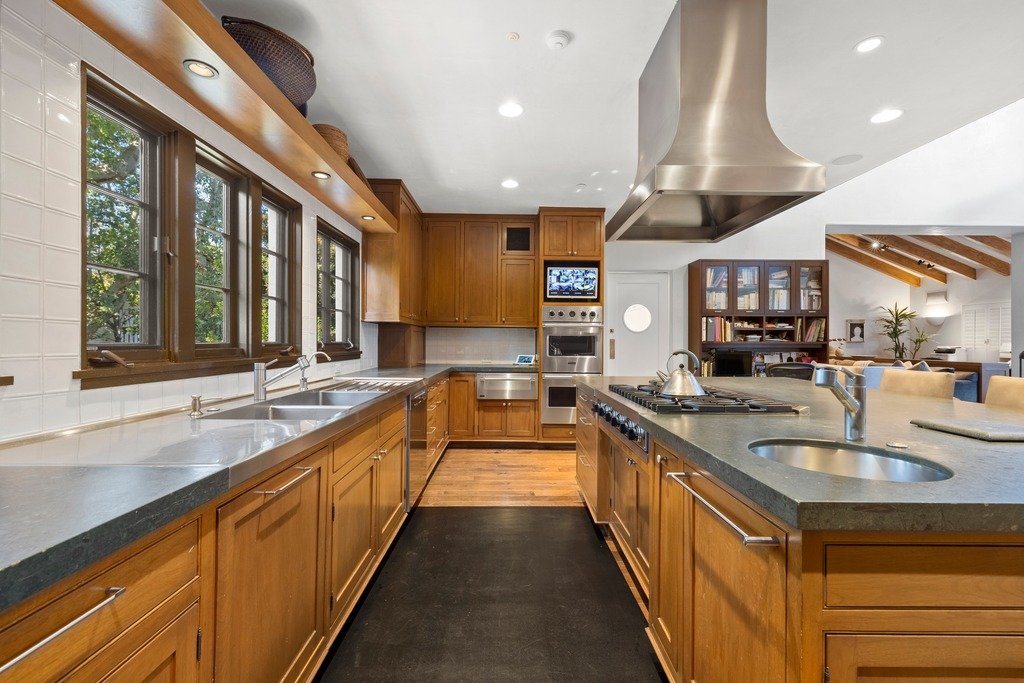 Spacious luxury kitchen in Brentwood Park with custom wood cabinetry, dark stone countertops, large island, stainless steel range with hood, built-in ovens, and multiple windows providing natural light.