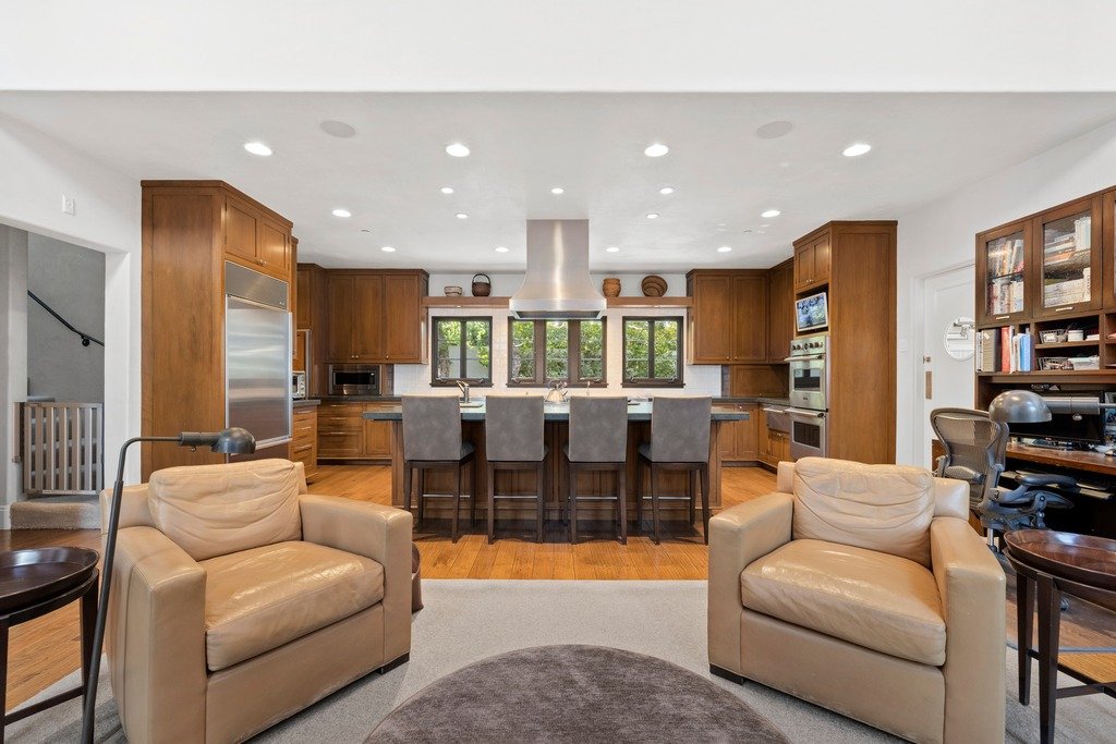 Open-concept living area with two light brown armchairs, a round dark ottoman, and side tables, flowing into a modern kitchen with wooden cabinetry, stainless steel appliances, and an island with four grey bar stools, plus a home office with built-in bookshelves visible on the right.