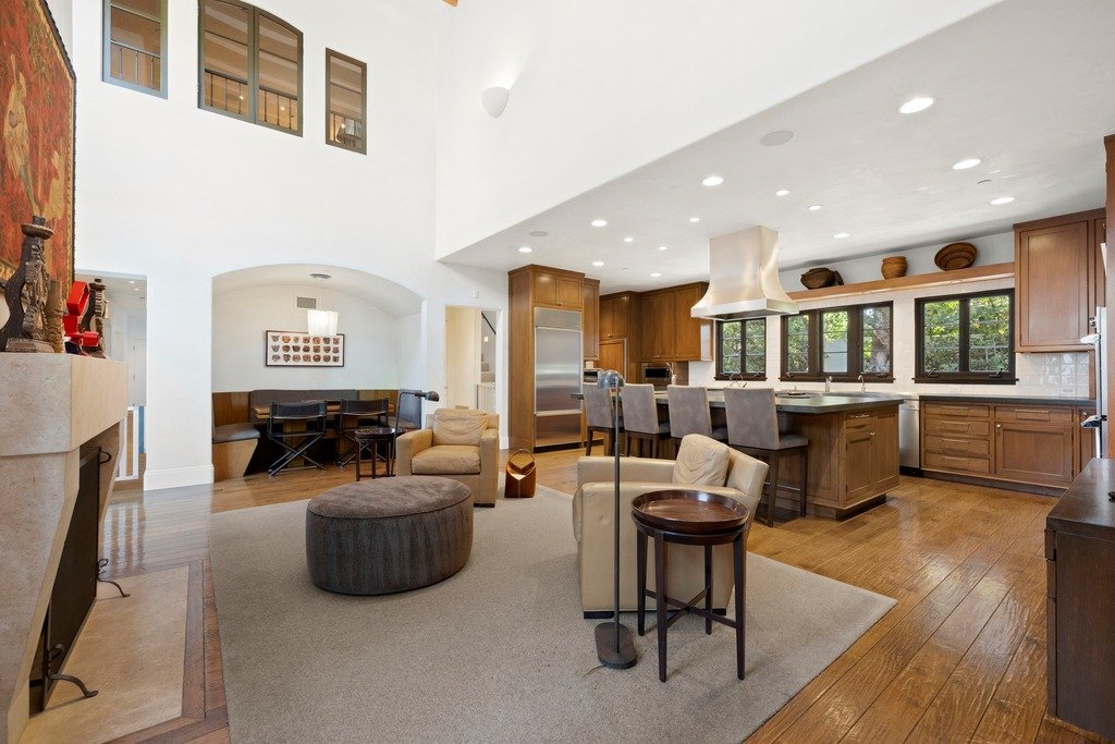 Spacious living room with exposed wooden ceiling beams, an intricate patterned area rug, modern and rustic furniture including a sofa, armchairs, and wooden coffee table, with large windows opening to a dining area and outdoor view.