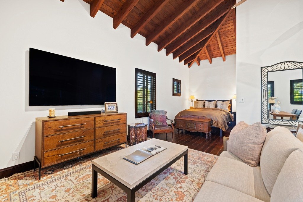 Cozy bedroom and living space with vaulted ceiling and exposed beams, featuring a bed with dark wood frame, sofa, flat-screen TV, wooden dresser, coffee table, window with shutters, and decorative mirror.