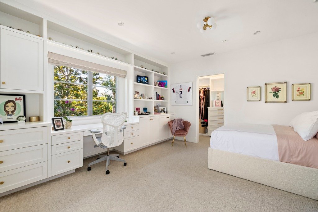 Bright bedroom with white built-in desk and shelves, window view of trees, white office chair, pink accent chair, bed with white and pink bedding, and walk-in closet.