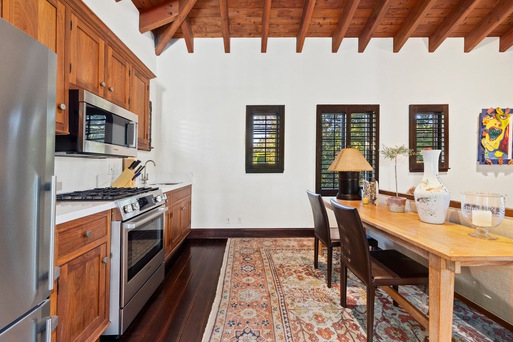 Rustic kitchen and dining area with warm wood cabinetry, ceiling beams, stainless steel appliances, patterned rug, wooden table, and shuttered windows.