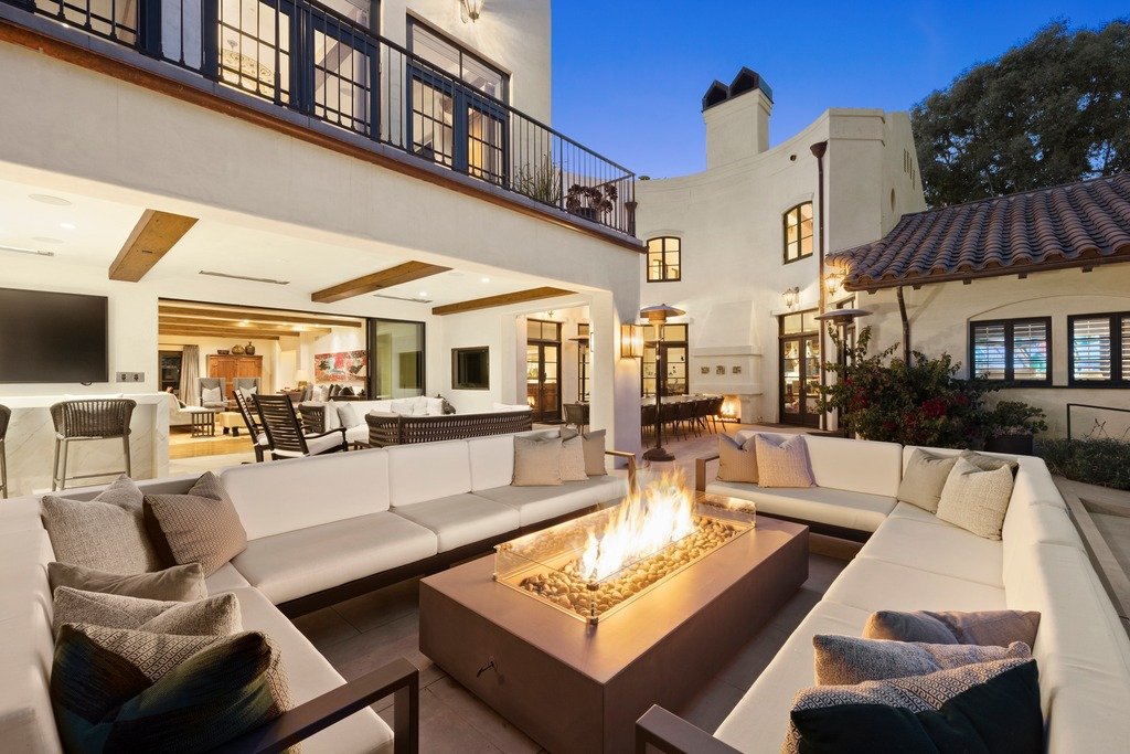 Luxurious outdoor living area at dusk with a U-shaped white sofa surrounding a glowing fire pit, a nearby bar area, and a house with balconies in the background.